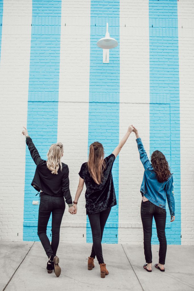 3 girls with their backs to the camera standing in front of a stripy blue and white wall, lifting their arms in the air