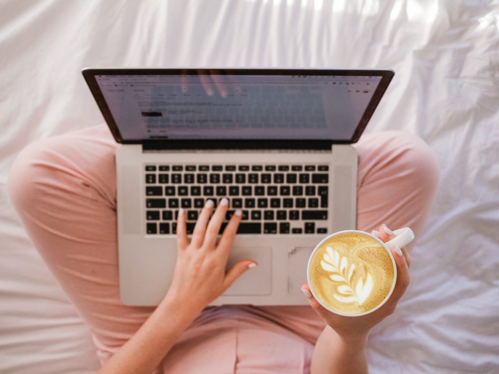 a capuccino and a laptop on a girl's knee as she sits in bed